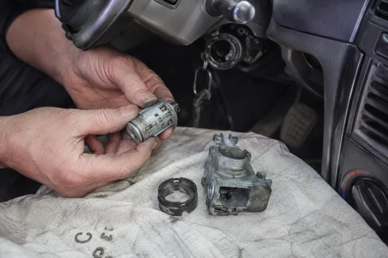 Hands holding a removed ignition cylinder beside a car steering column, with the housing and small parts on a cloth. — Ignition Lock Repair, Georgina